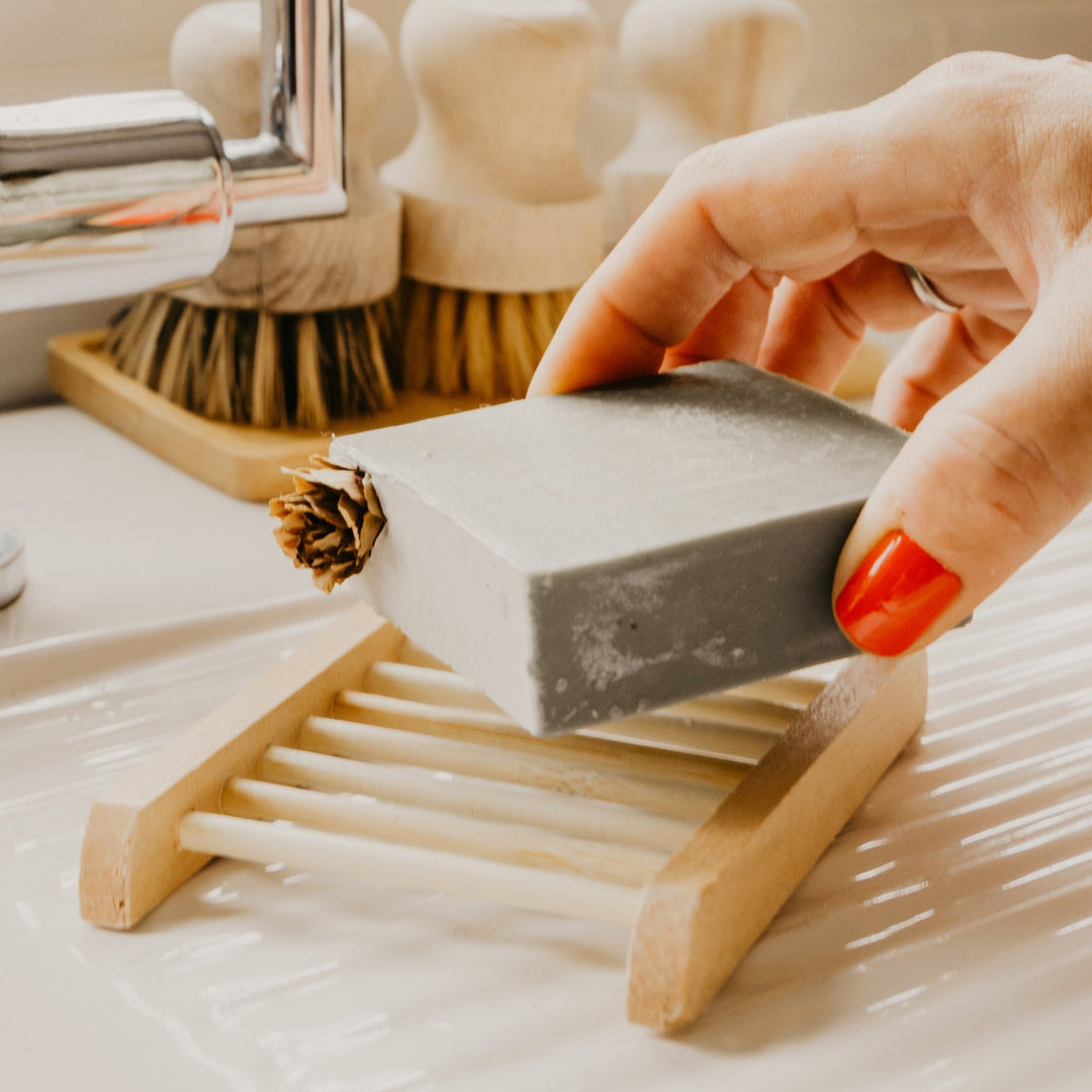 A soap bar over a small wooden BAMBOO soap dish in a bathroom sink, with soap brushes and a faucet blurred in the background.