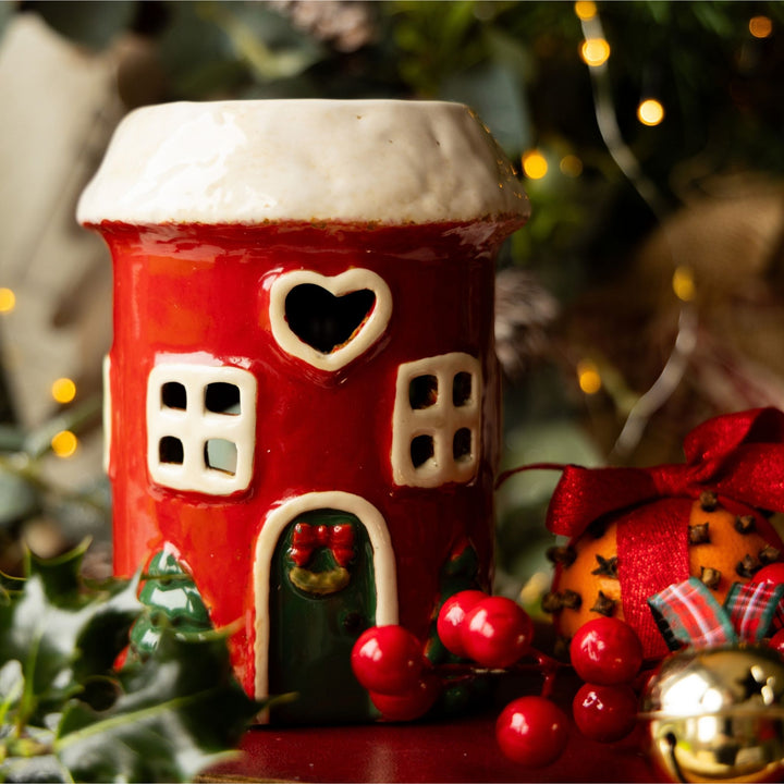 A red ceramic gingerbread house container with a white snowy roof, decorated with a heart-shaped window, square windows, and a green door with a red bow, styled with holiday greenery and ornaments.