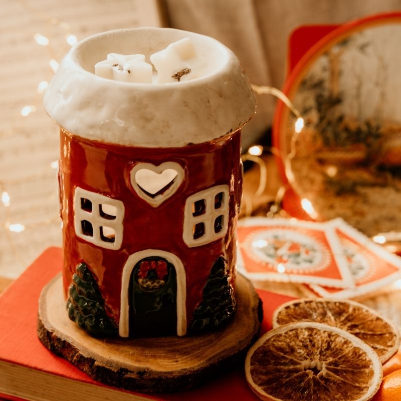 A red ceramic gingerbread house container with a white snowy roof, decorated with a heart-shaped window, square windows, and a green door with a red bow, styled with holiday greenery and ornaments.