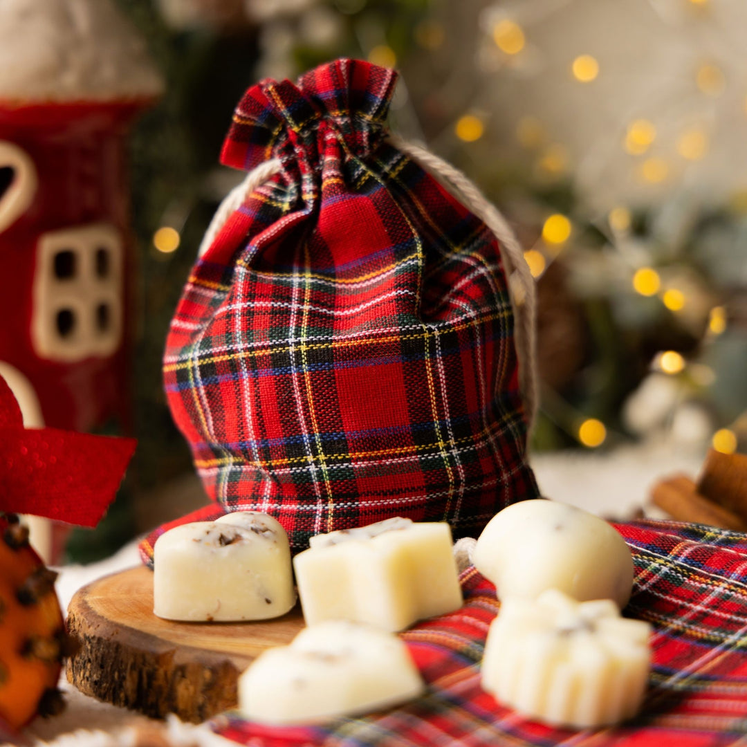 A red plaid tartan drawstring gift bag filled with cream-colored wax melts, styled on a wooden slice with more wax melts and a festive holiday background.