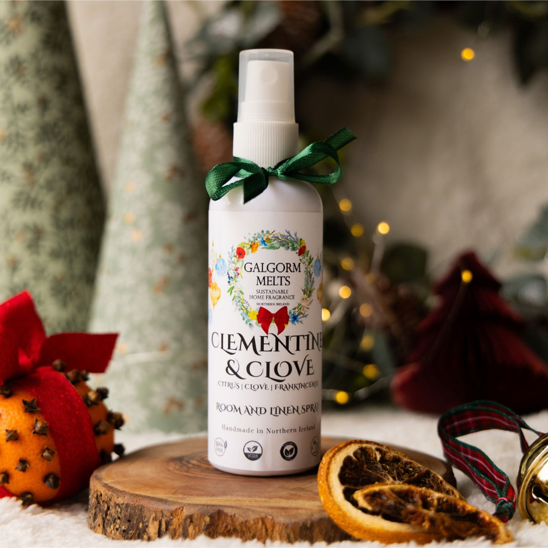 A bottle of Galgorm Melts 'Clementine & Clove' room and room spray stands on a wooden slice coaster. The white spray bottle has a green ribbon tied around its neck and is surrounded by festive Christmas props including a dried orange slice, a red ornament, and soft holiday lights in the background.