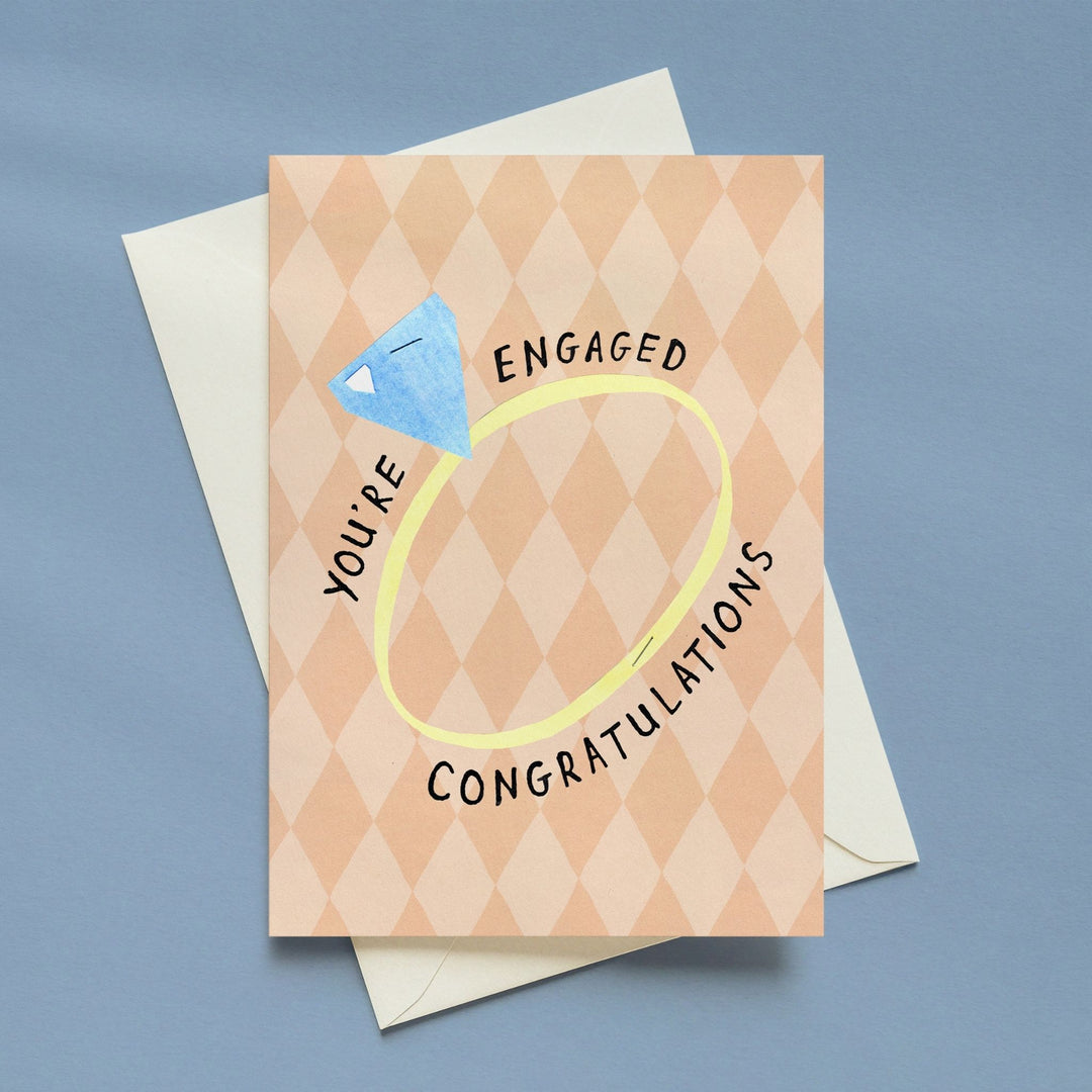 A square congratulations card with a peach and beige harlequin pattern, featuring a blue diamond ring graphic and the words &