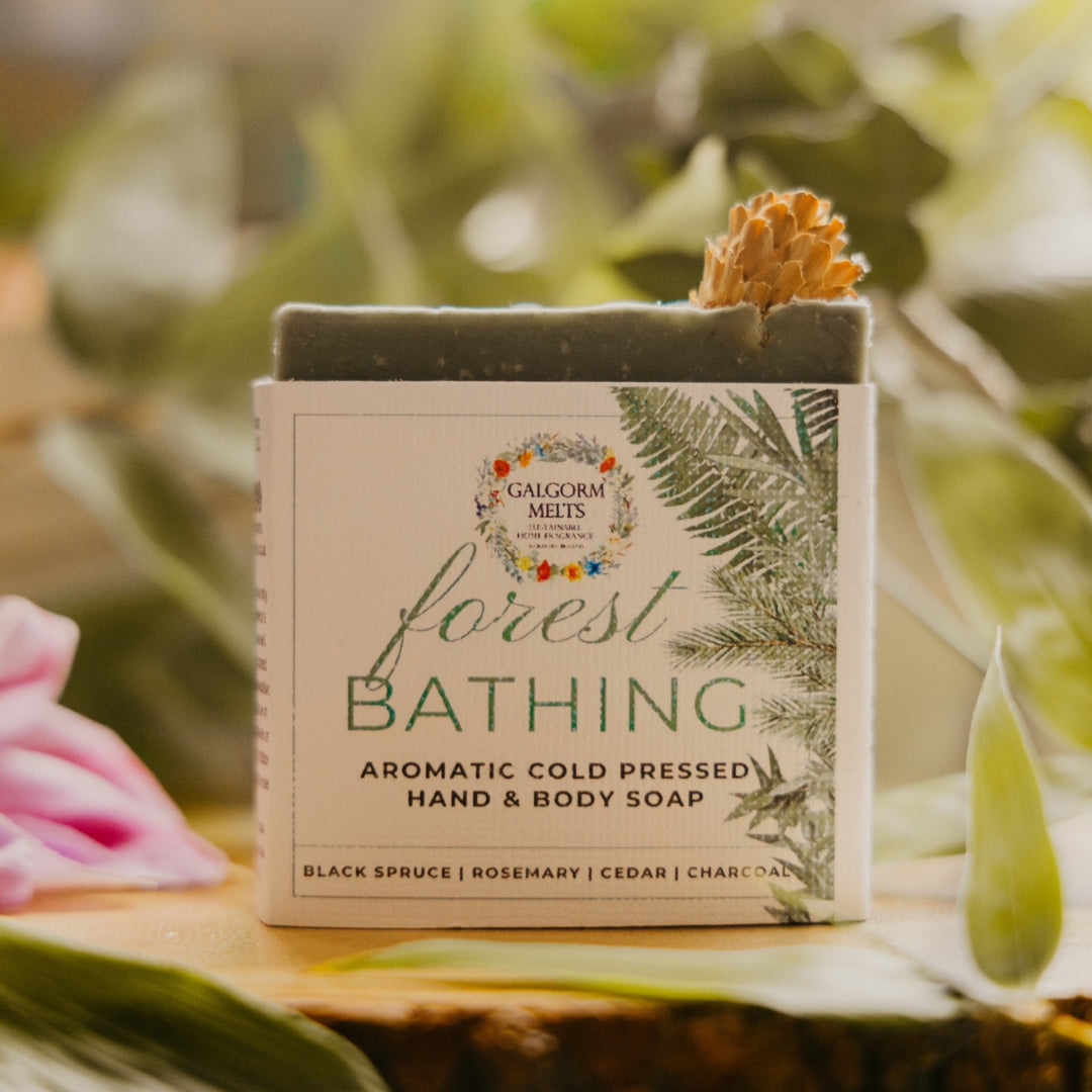 A rectangular bar of hand and body soap labeled 'forest BATHING' by Galgorm Melts, placed on a leafy surface with blurred green foliage and a pink flower in the background.