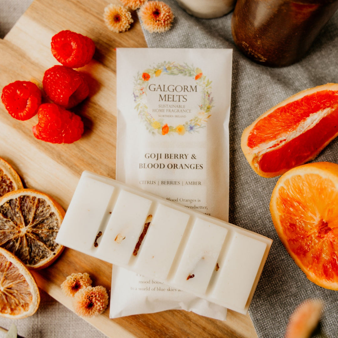 A BEST SELLING SOY WAX MELT BY GALGORM MELTS, GOJI BERRY AND BLOOD ORANGES IS FRESH AND FRUITY IN FRAGRANCE. SITTING ON A WOODEN PLINTH SURROUNDED BY FRUITS.