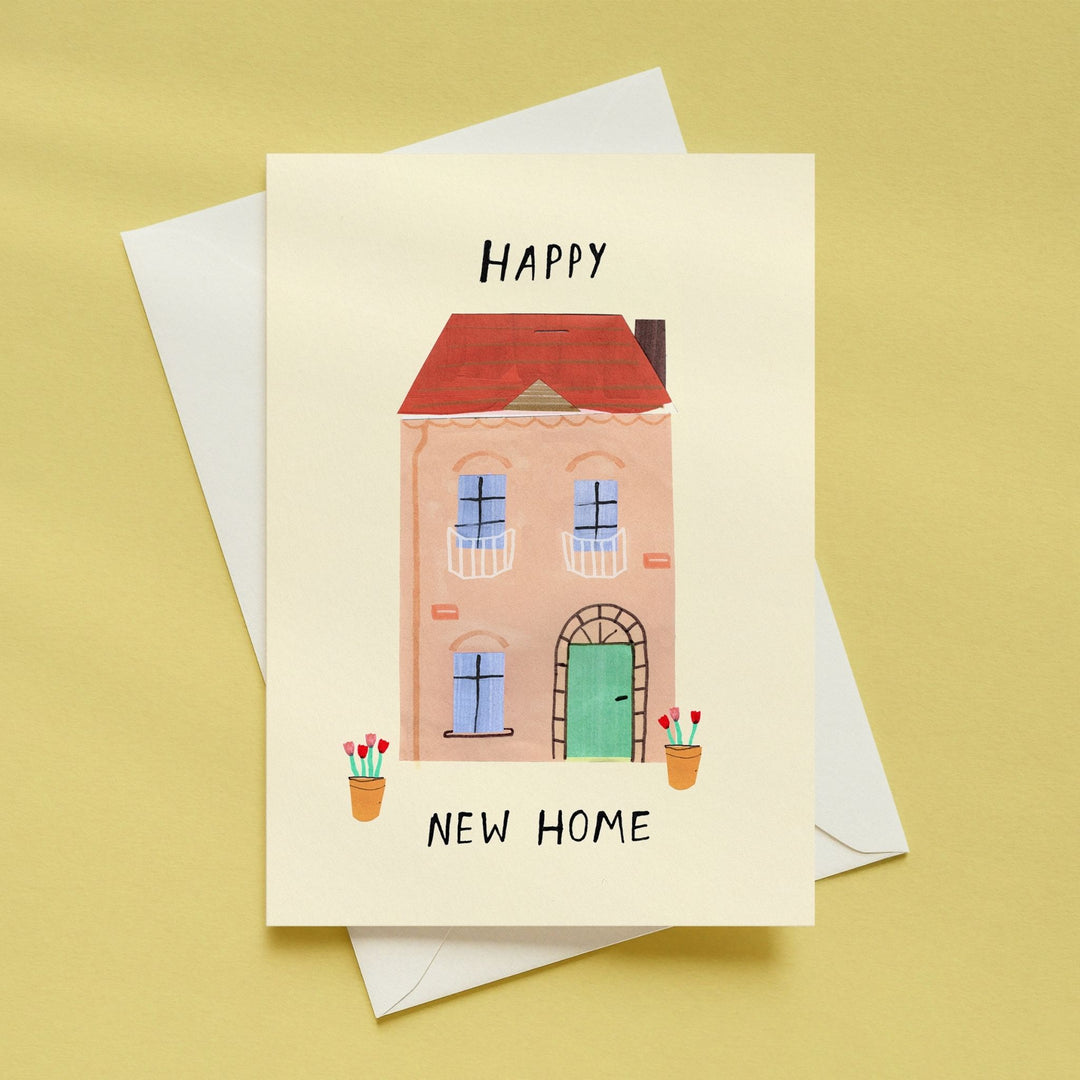 A cream-colored greeting card with a hand-drawn illustration of a pink house, red roof, green door, and potted flowers, with the words &