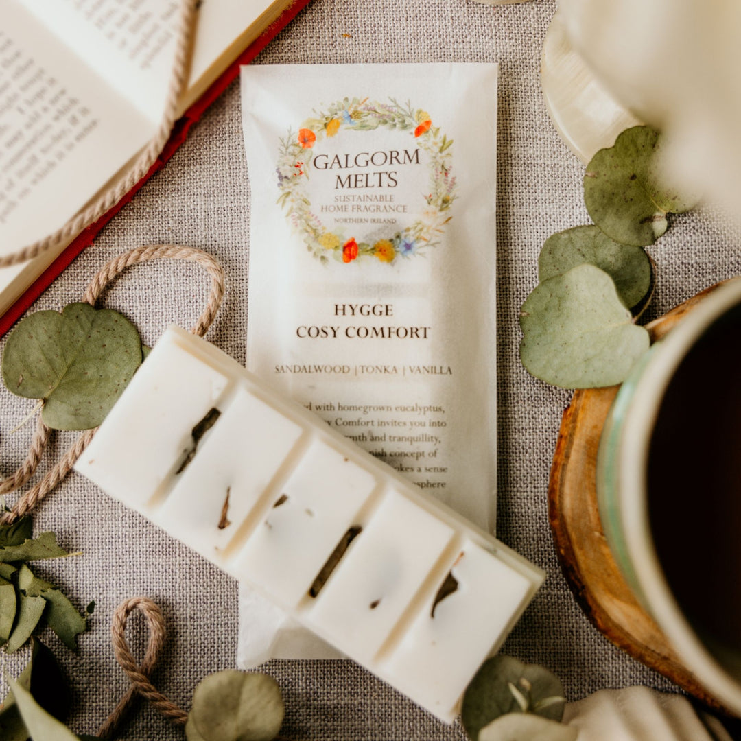 A package of Galgorm Melts Hygge Cosy Comfort home fragrance is shown with several white wax melts in a metal tin placed on a linen surface, surrounded by eucalyptus leaves, rope, an open book, and a mug.