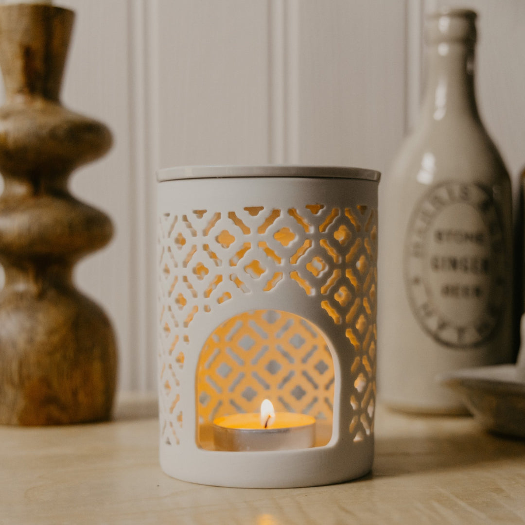 A white ceramic tea light holder with geometric cutout patterns, containing a lit tea light candle, set on a wooden surface with a warm, cozy background.