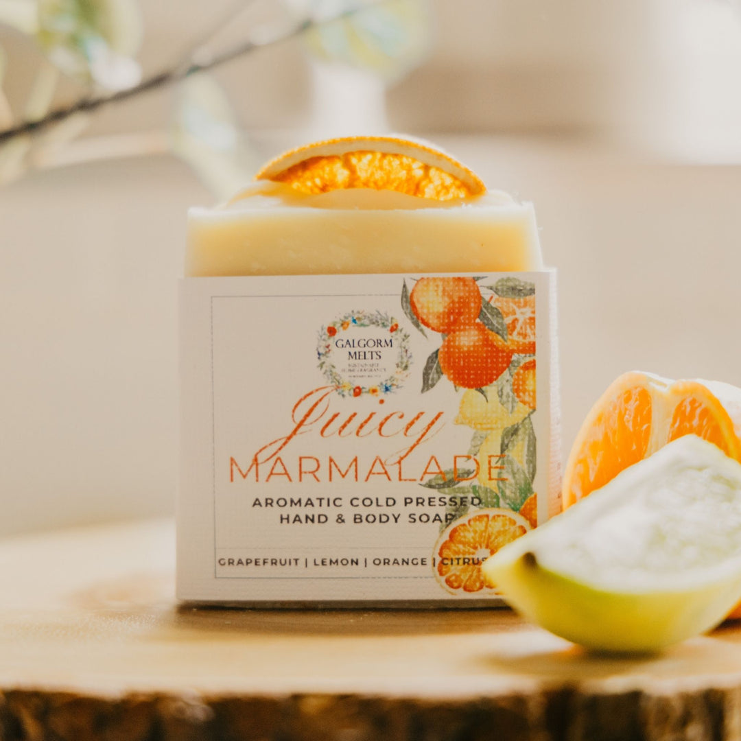 A bar of hand and body soap labeled 'Juicy Marmalade' by Galgorm Melts, styled on a rustic wood slice with orange and grapefruit slices in a warm, softly blurred setting.