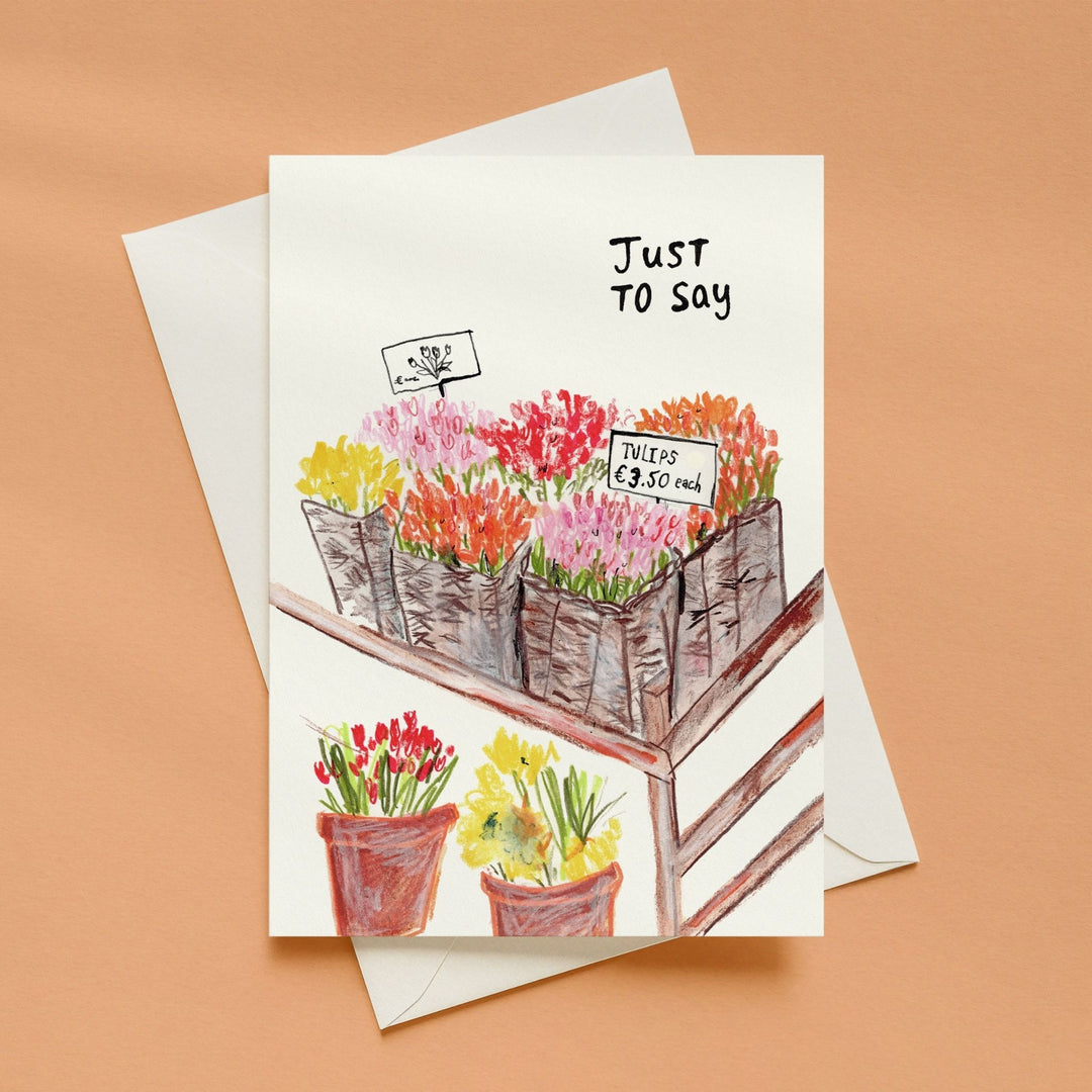 A white greeting card with a watercolor illustration of flower boxes and potted flowers, with the handwritten text 'Just to say' at the top, shown on a peach background with a white envelope behind it.