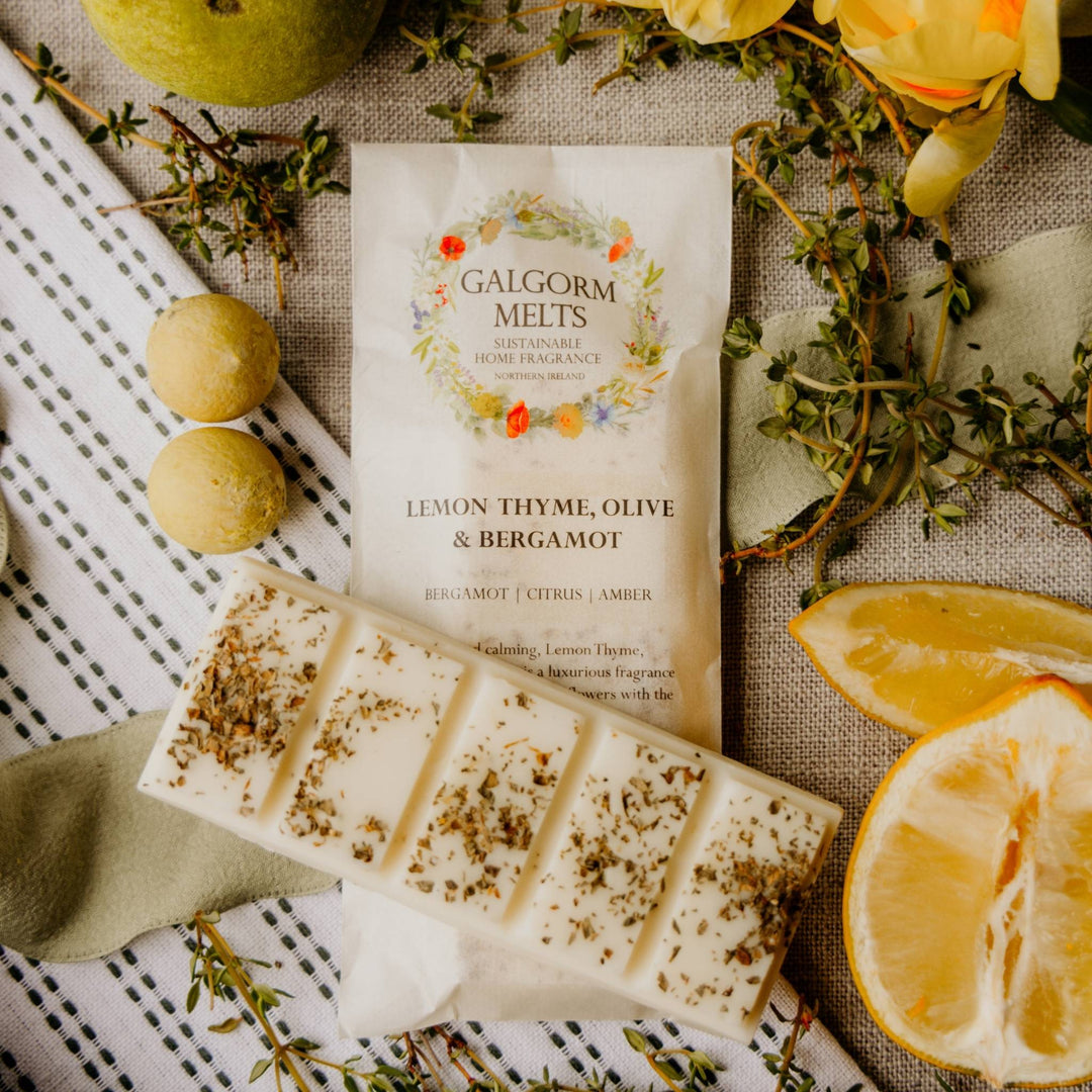 A package of Galgorm Melts home fragrance in scent, Lemon Thyme, Olive & Bergamot, styled in a flat lay with actual lemon slices, bergamot fruit, and fresh herbs on a linen background.