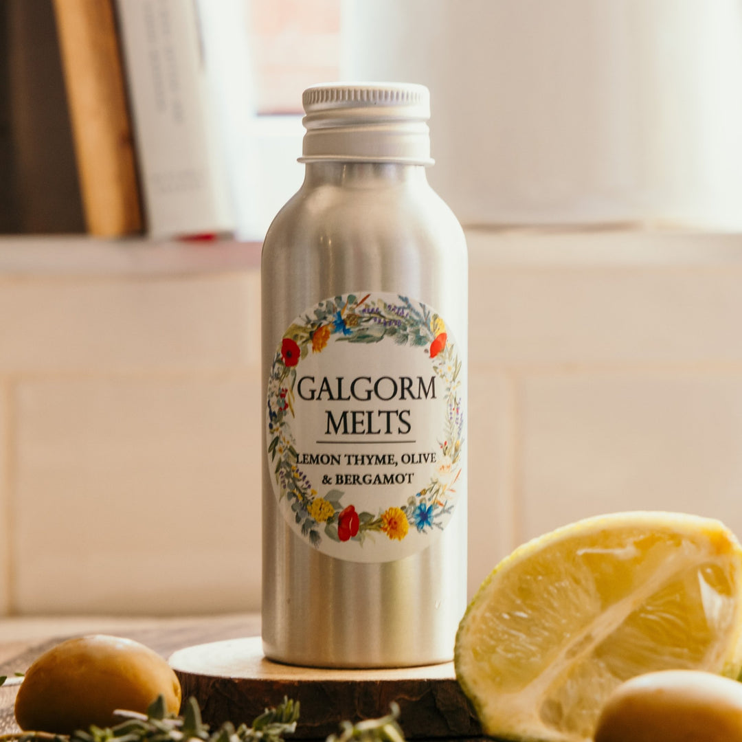 A silver metal bottle of GALGORM MELTS 'Lemon Thyme, Olive & Bergamot' reed diffuser refill sitting on a wooden slice. The bottle is decorated with a colorful floral wreath label.