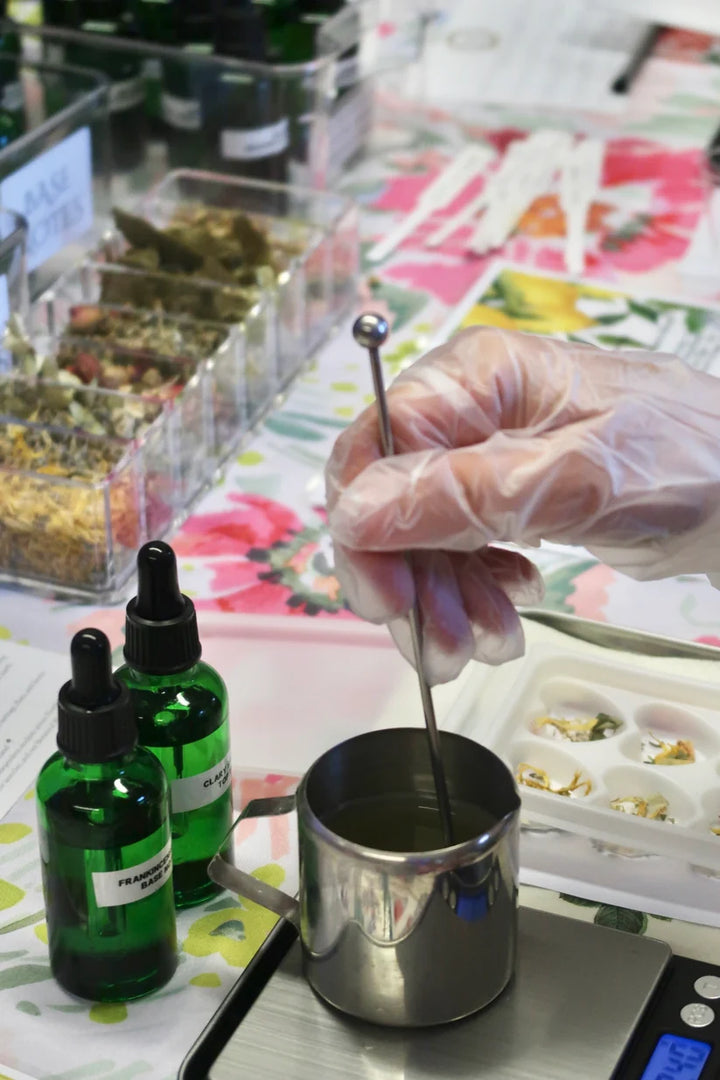 A close-up of a hand wearing a clear plastic glove stirring a small stainless steel pot on a digital scale. Two green glass dropper bottles with black caps sit nearby on a floral table surface, with ingredient containers and other workshop items blurred in the background.