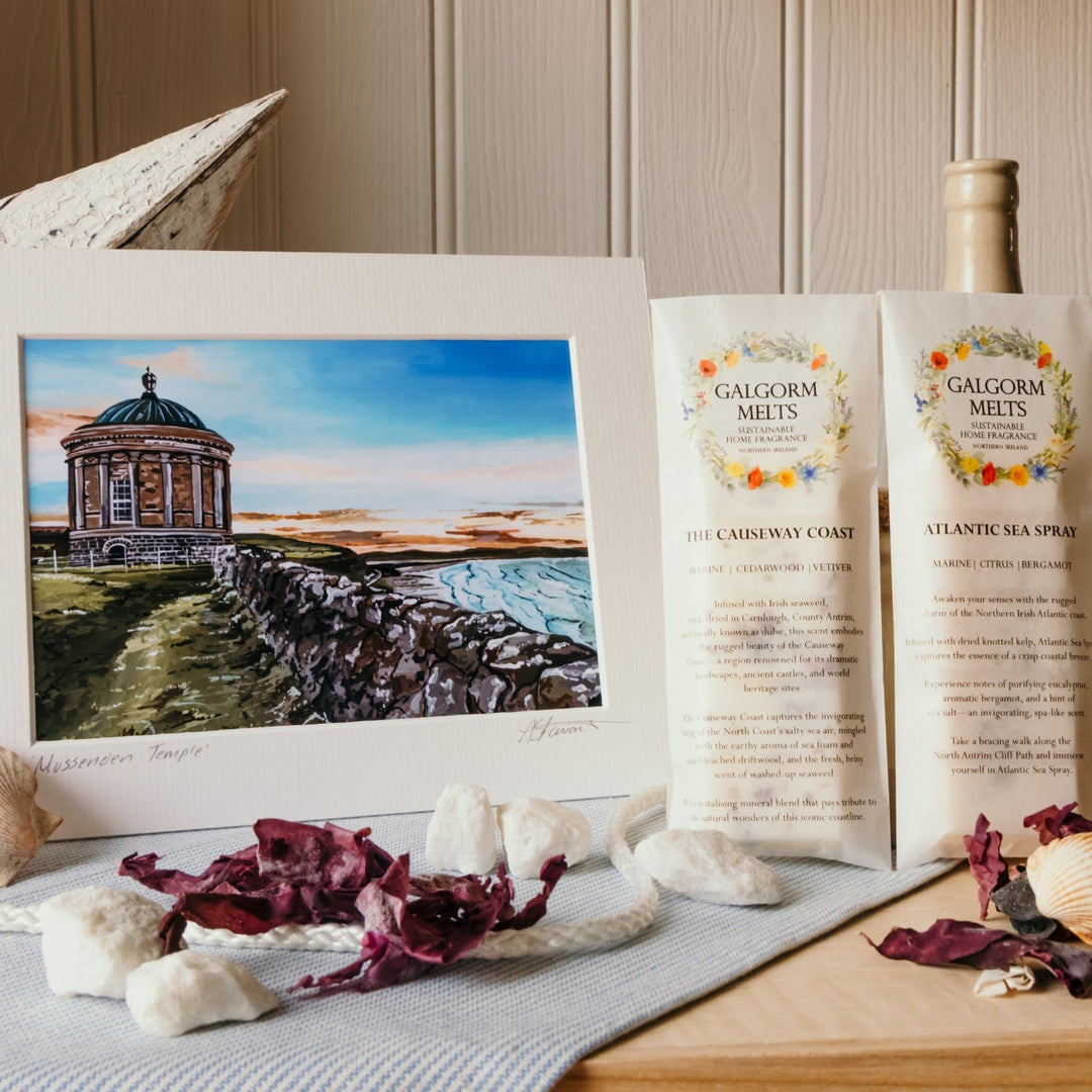 A styled product display featuring a framed watercolor painting of a coastal building and sea wall, alongside two tubes of Galgorm Melts and a bottle of Atlantic Sea Spray on a tabletop with dried seaweed and shells.