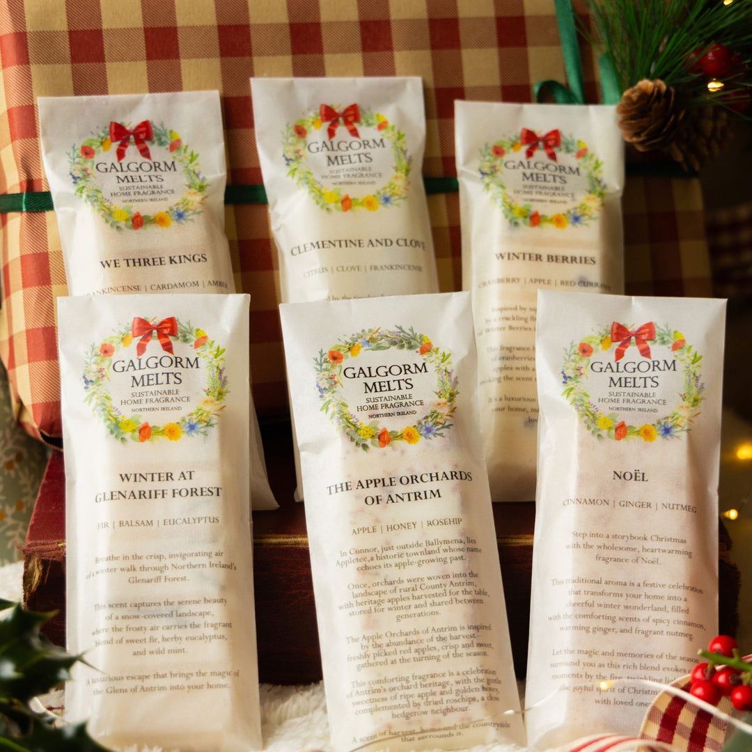 Six bags of Galgorm Melts soy wax melts arranged on a white surface in front of a red and green plaid gift bag, with Christmas decorations including pine branches and lights in the background.