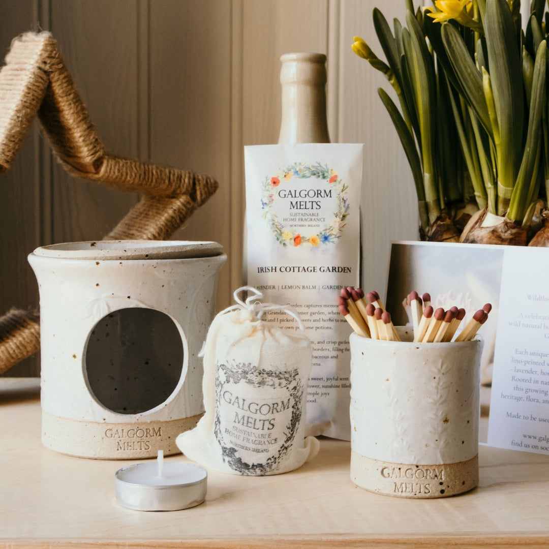 A styled product display of Galgorm Melts products on a wooden surface, including a ceramic wax warmer, a small fabric bag of tealights, a match holder filled with matches, and a packet of Irish Cottage Garden melts, styled with daffodils and a wicker basket.