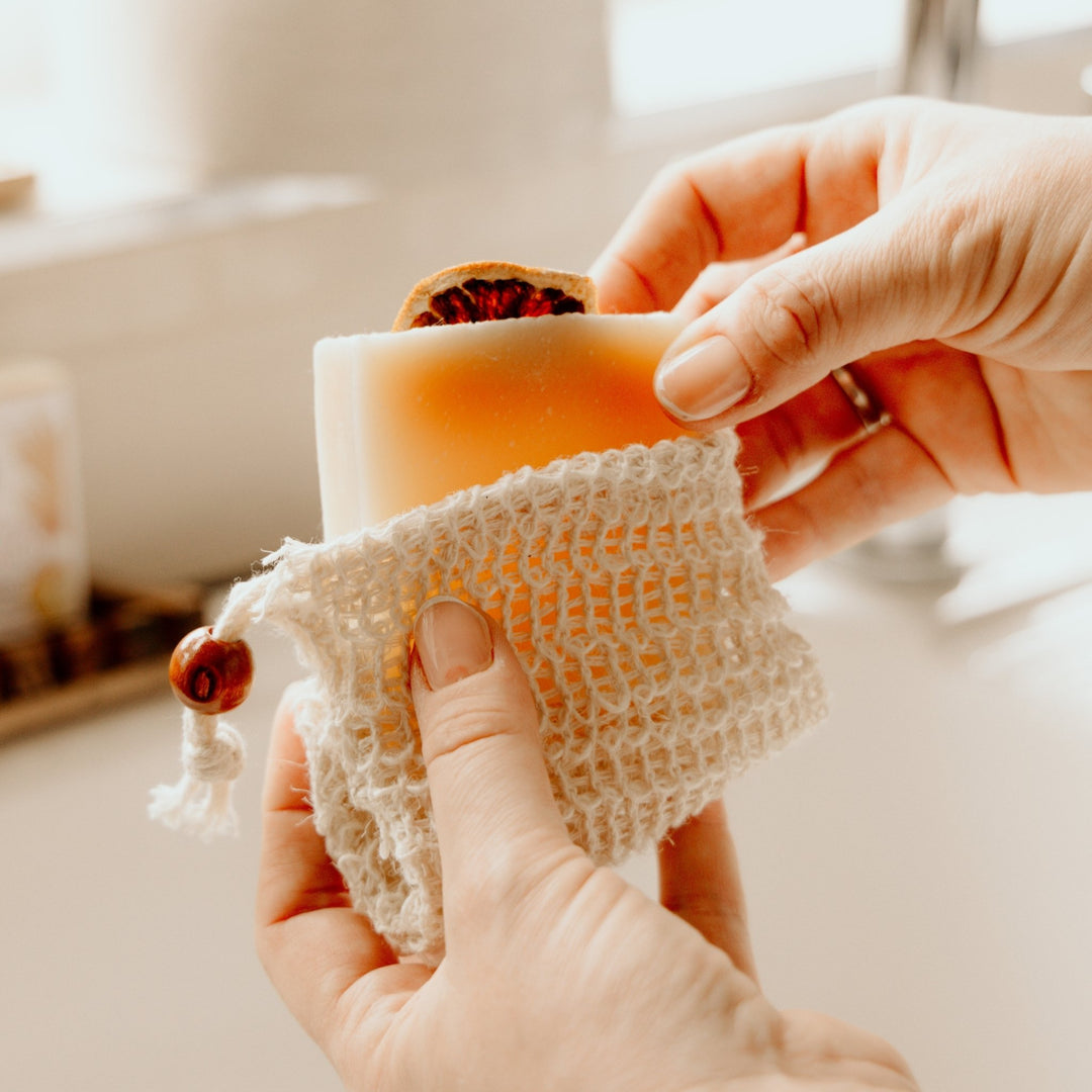 A close-up of hands holding a handmade soap bar wrapped in a cream-colored cotton mesh soap bag, with a dried rose petal garnish, against a softly blurred bathroom background.