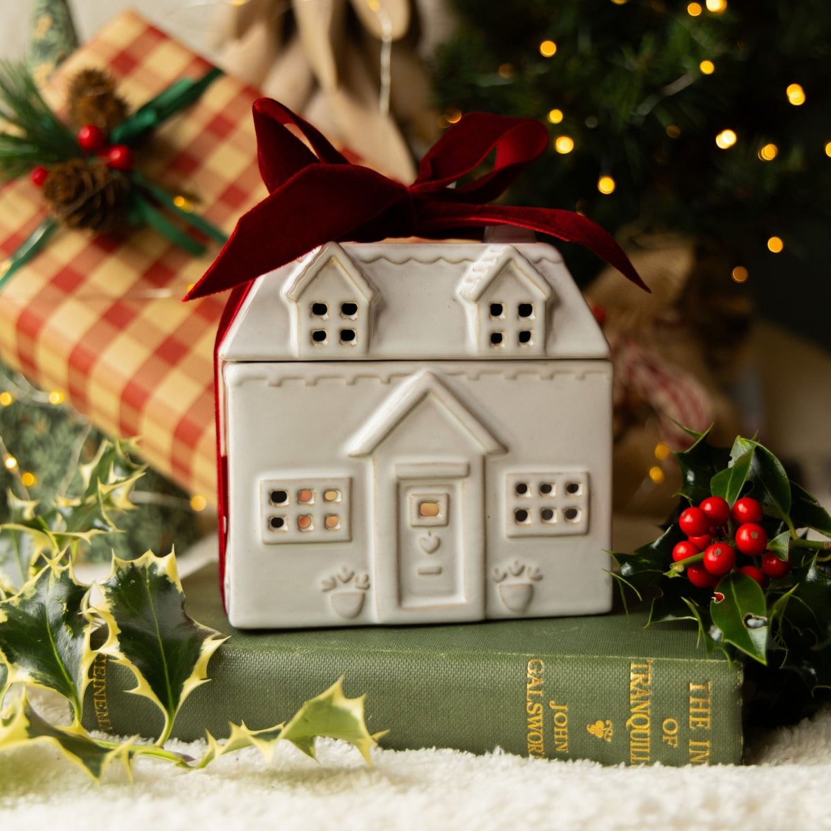 A small white ceramic house-shaped container or box with a dark red ribbon bow, styled on a green book with holly leaves and red berries, with a wrapped gift and blurred Christmas lights in the background.