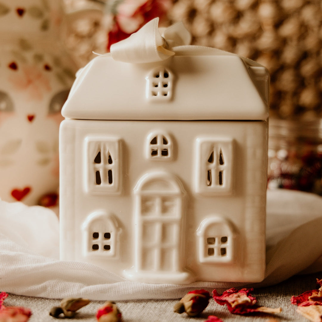 SANDYMOUNT WAX WARMER. A small white ceramic house-shaped container or ornament with cutout windows and a bow on the roof, photographed in a softly styled setting with warm blurred background elements.