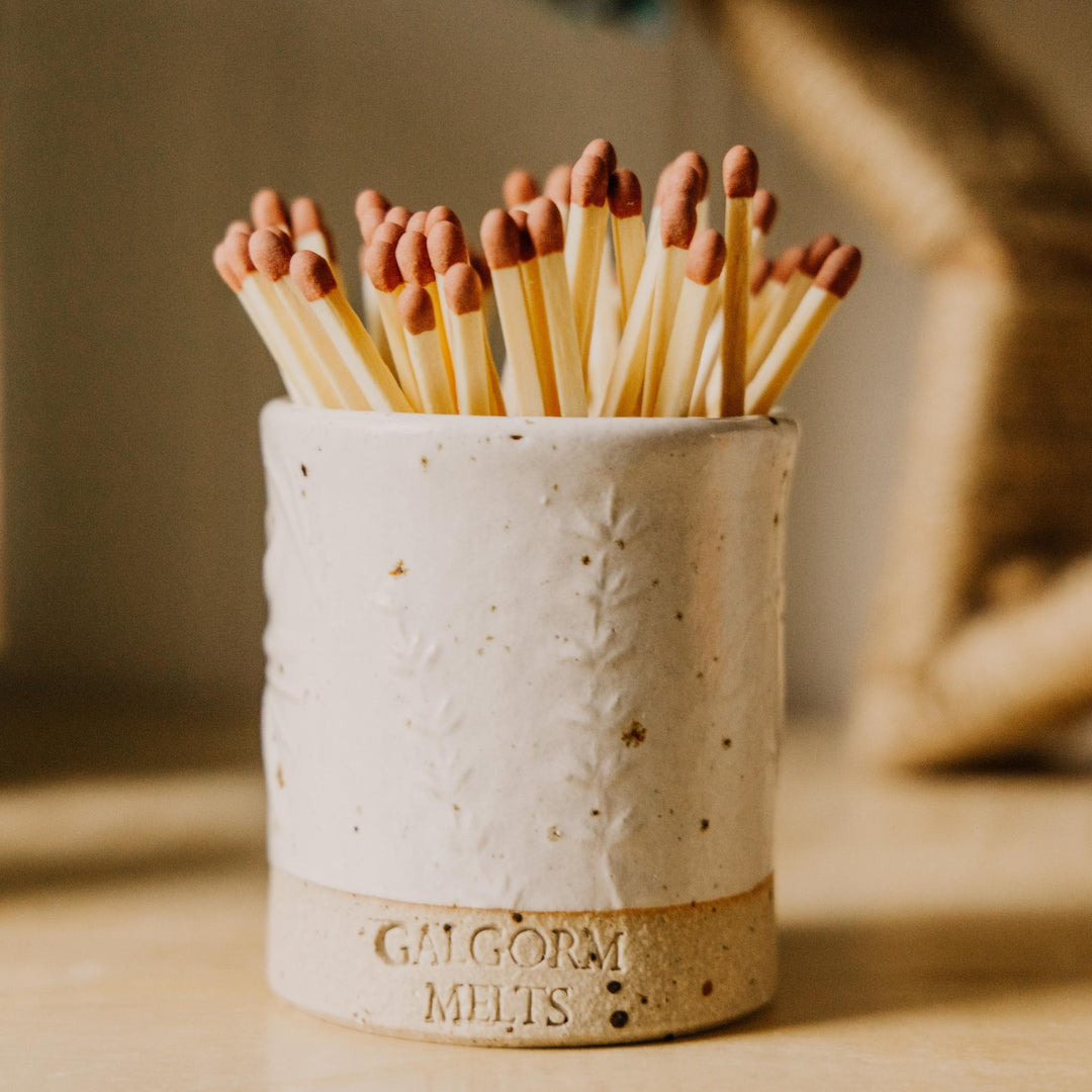 A handmade ceramic match holder with the words 'GALGORM MELTS' embossed on the base, filled with wooden matches with copper-colored tips, set against a warm, softly blurred background.