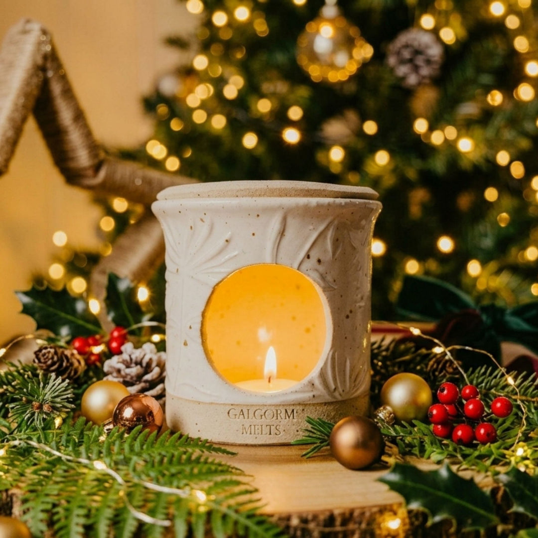 A ceramic holiday candle holder by Calgornie Melts with a lit candle inside, surrounded by festive greenery, pinecones, and a blurred Christmas tree background.