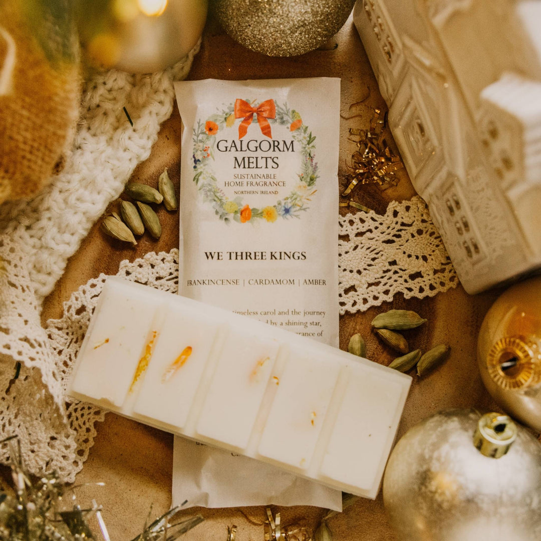 A packaged wax melt product labeled 'Galgorm Melts' and 'We Three Kings' is displayed with a partially melted wax bar in a styled holiday flat-lay. The scene includes lace, gold ornaments, cardamom pods, and warm ambient lighting.