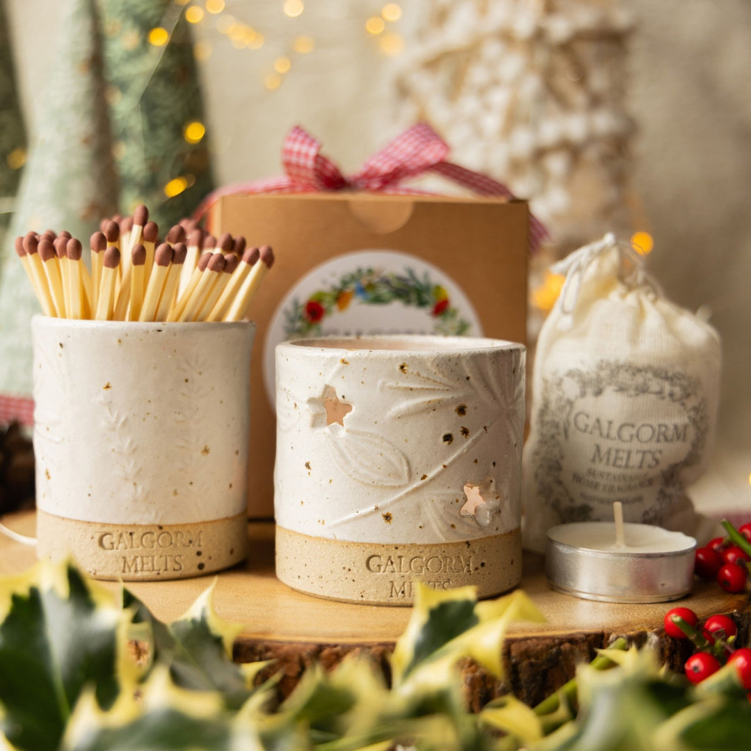 A styled holiday gift set featuring a ceramic match holder filled with matches, a matching ceramic scented candle holder, a bag of Galgorm Melts, and a small tea light candle, arranged on a wood slice with holly and warm blurred lights in the background.