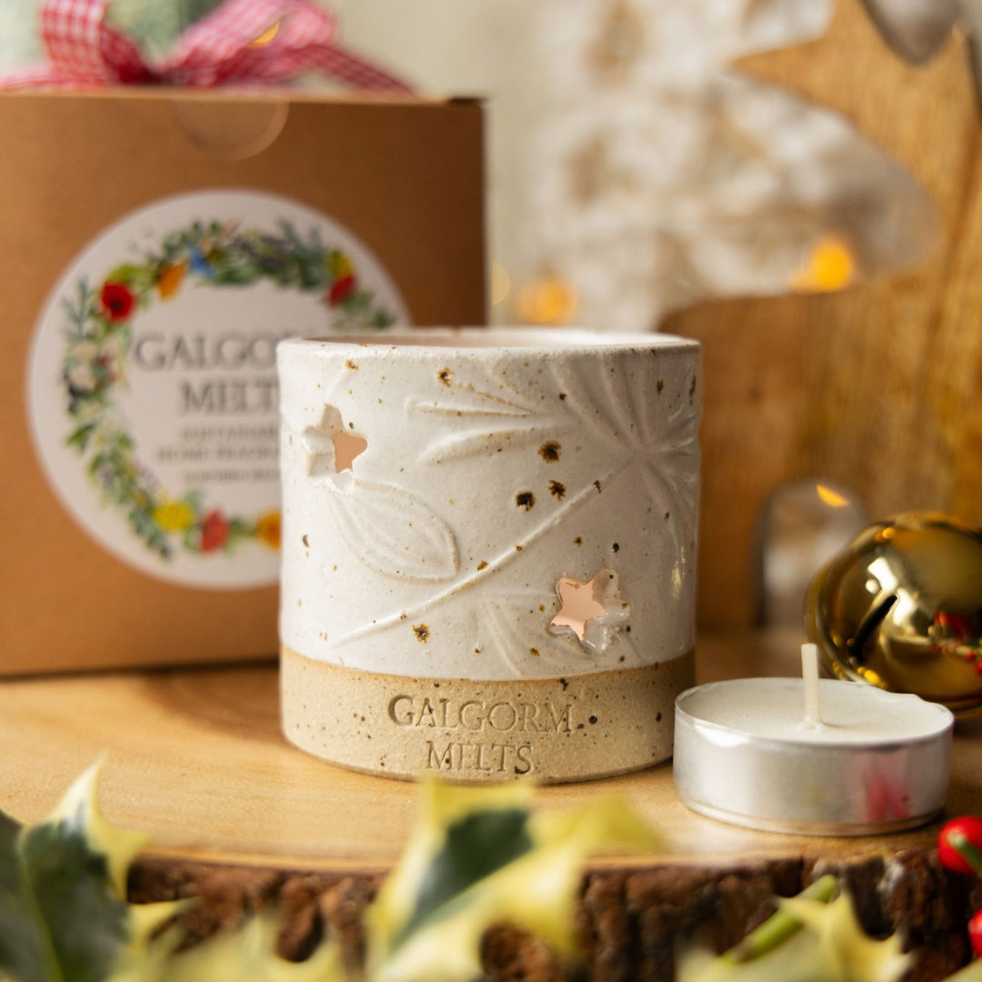 A ceramic holiday candle holder with star-shaped cutouts sits on a wooden surface, with its branded box blurred in the background and a small tea light candle beside it.