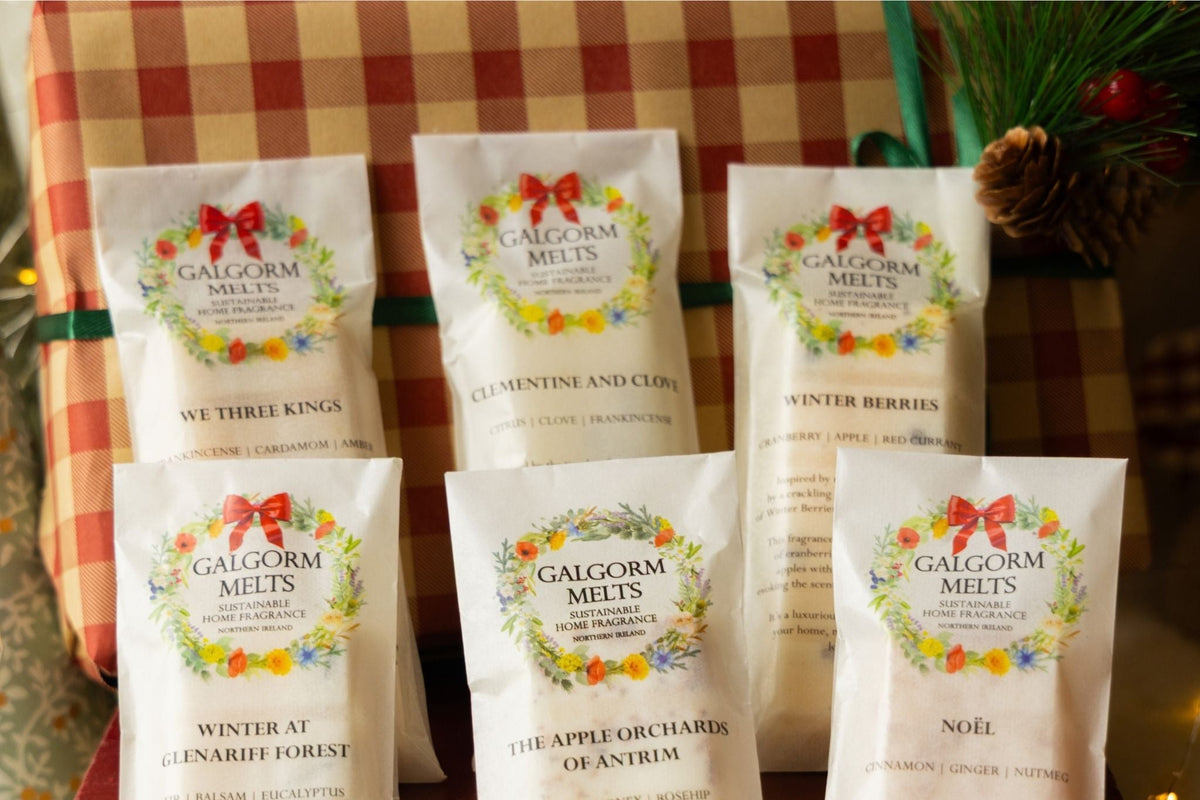 Six bags of Galgorm Melts Christmas melts arranged in a gift basket with a red and white checkered wrapping paper background, decorated with pinecones, evergreen branches, and red berries.